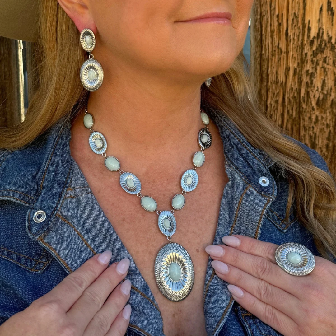 A woman wears The Kathryn Drop Earrings as part of a matching silver-toned set featuring oval, light-colored stones, including a necklace with a large central pendant and a ring.