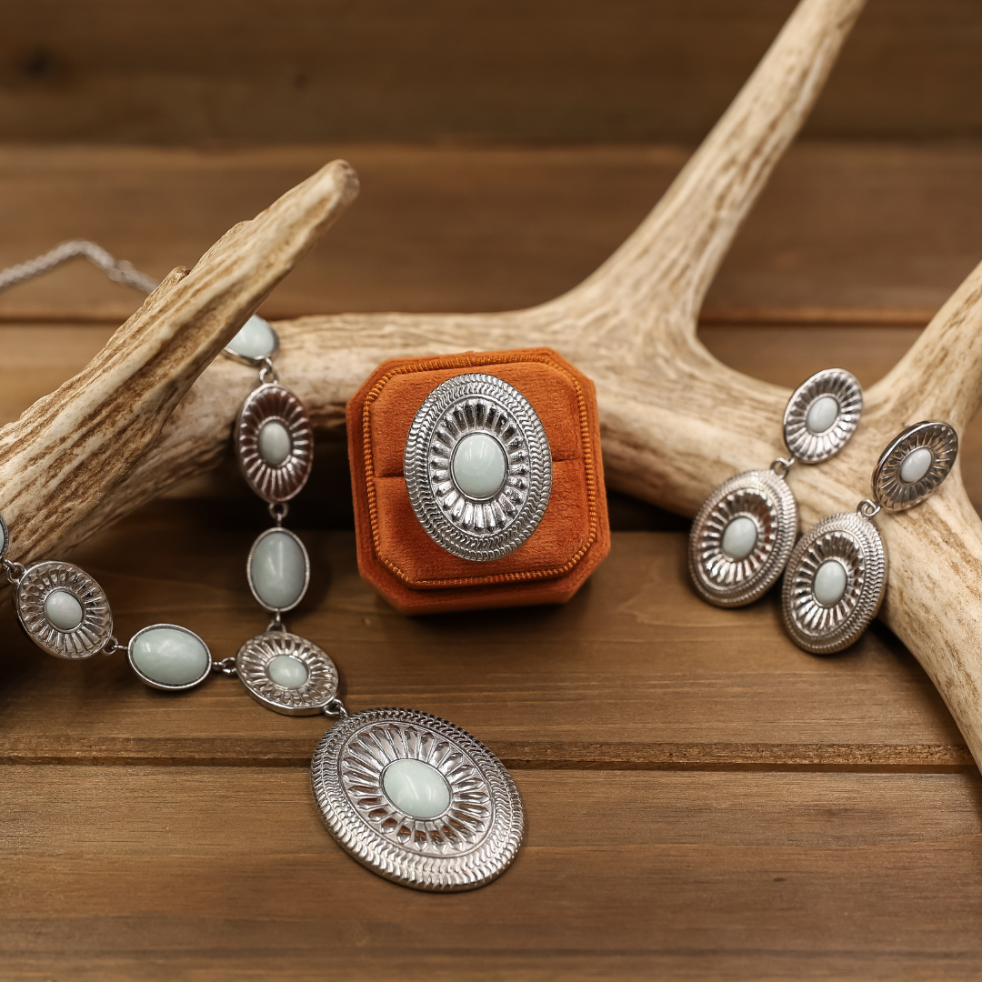 The Kathryn silver Western jewelry set includes an oval light blue statement necklace, ring, and drop earrings, arranged around an orange velvet ring box on wooden antlers.