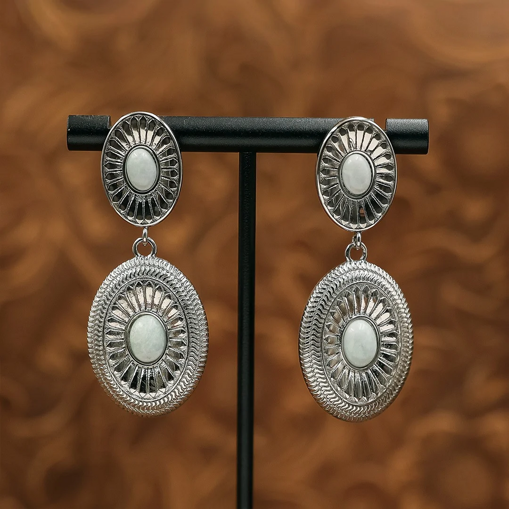 The Kathryn Drop Earrings in silver-tone, featuring two oval, etched-metal discs with white stone centers, displayed on a black stand.