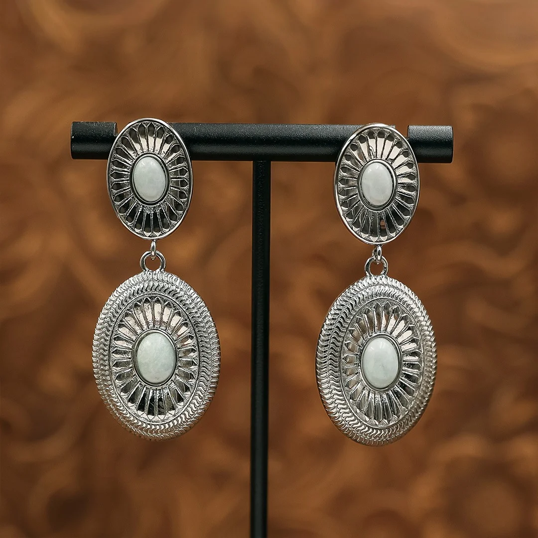 The Kathryn Drop Earrings in silver-tone, featuring two oval, etched-metal discs with white stone centers, displayed on a black stand.