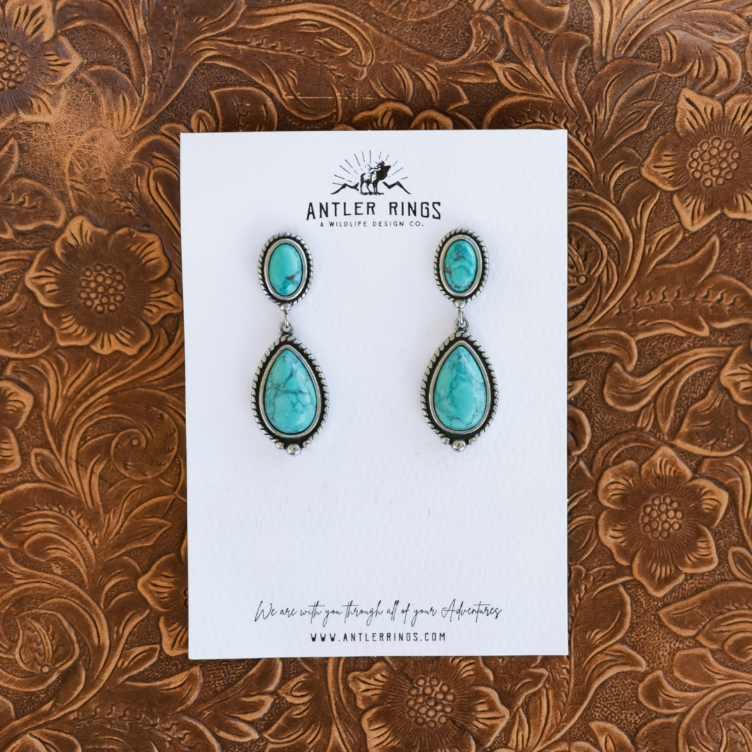 The Kay Drop Earrings, featuring oval and teardrop-shaped turquoise stones set in silver, displayed on a branded white backing card.