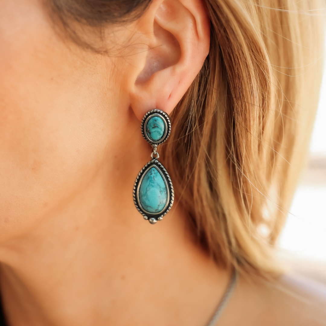 The Kay Drop Earrings feature an oval turquoise stone topped by a smaller round turquoise stone in silver settings, shown worn on an ear.