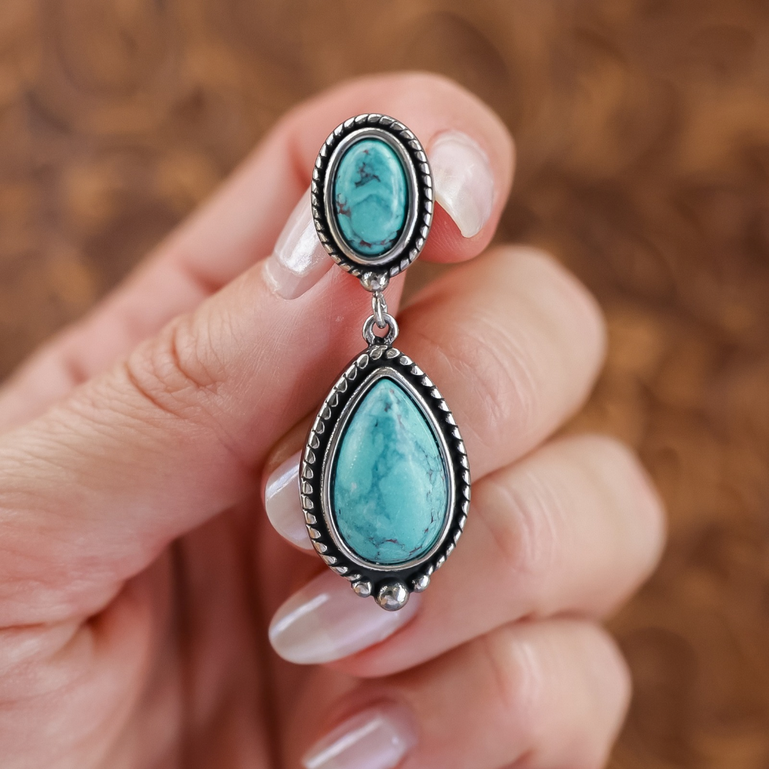 A hand holds a pair of The Kay Drop Earrings, featuring a small oval turquoise stone above a larger teardrop-shaped turquoise stone set in silver.
