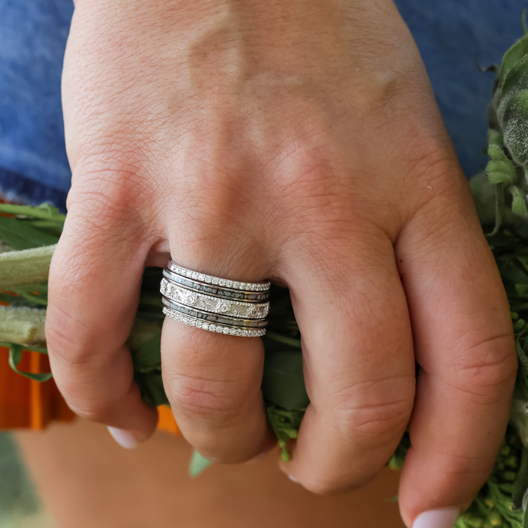 A close-up of a hand wearing The Kenai Florence CZ Stacker Set—several stacked silver bands with sparkling cubic zirconia accents—holding a bundle of leafy greens.
