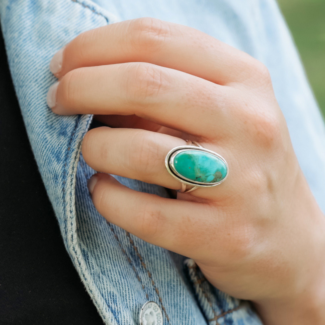 A hand wearing The Lainey, a bold Mohave turquoise statement ring, rests on a light blue denim jacket, with short, neatly manicured nails.