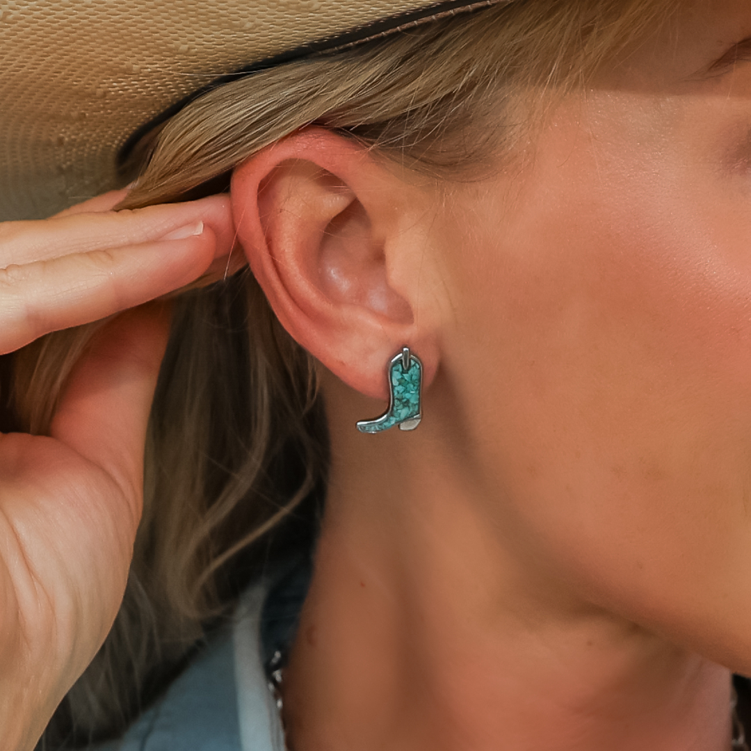 A person wearing The Boot Studs, turquoise and silver cowgirl boot earrings, touches their ear. The person has blonde hair and is wearing a light brown hat.