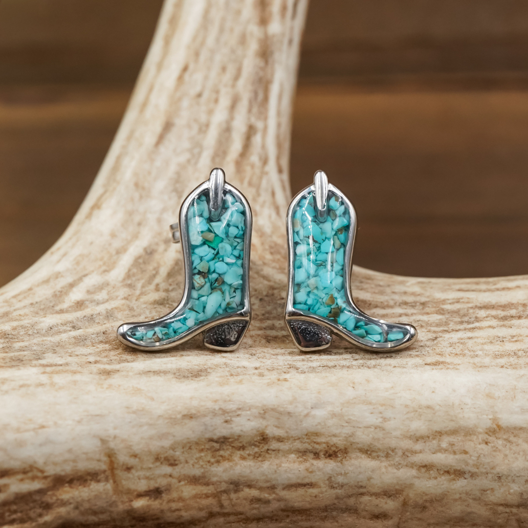 The Boot Studs—turquoise and silver cowgirl boot earrings—rest on a light tan antler against a softly blurred brown background. Each earring features petite turquoise stones set in silver, showcasing the unique charm of these boot-shaped stud earrings.