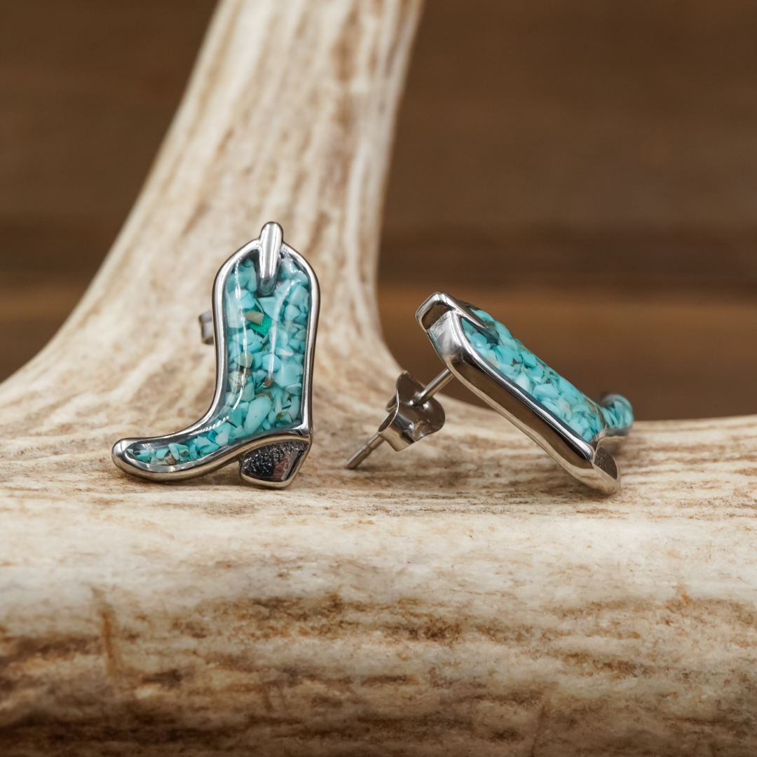 Two pairs of The Boot Studs, featuring turquoise and silver cowgirl boots, are displayed on a large antler. One earring stands upright while the other lies on its side, showcasing both the front and side views of their intricate design.
