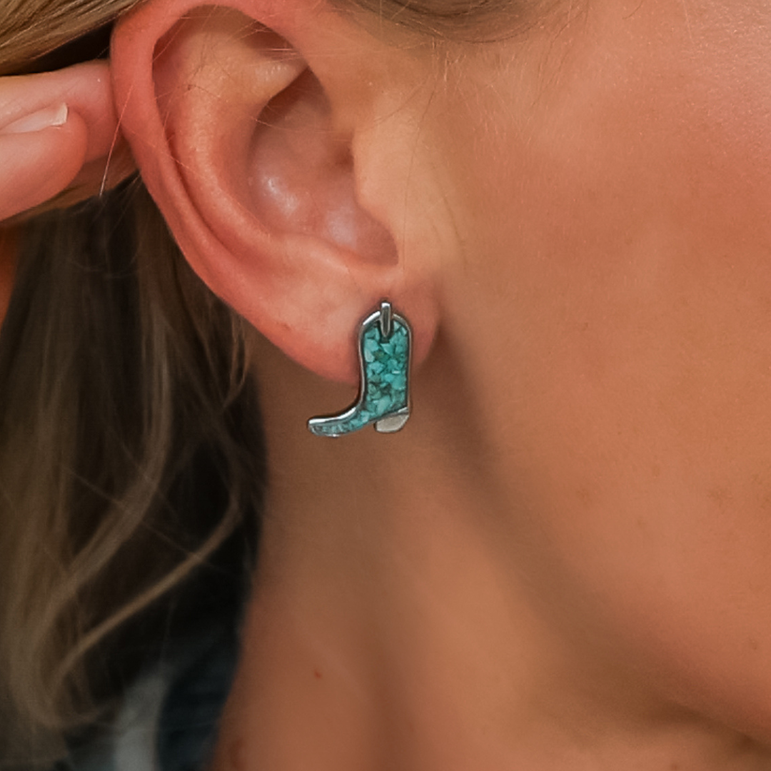A close-up of a person's ear wearing The Boot Studs, a turquoise cowgirl boot earring. The person has light skin and blond hair tucked behind the ear, with fingers gently touching the ear.