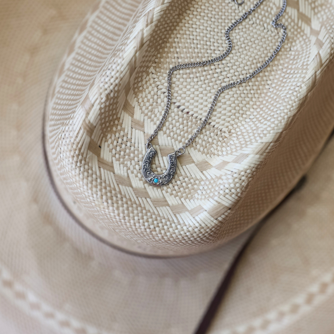 The Lucky Necklace, featuring a silver horseshoe and turquoise pendant, is draped across the brim of a woven beige cowboy hat.