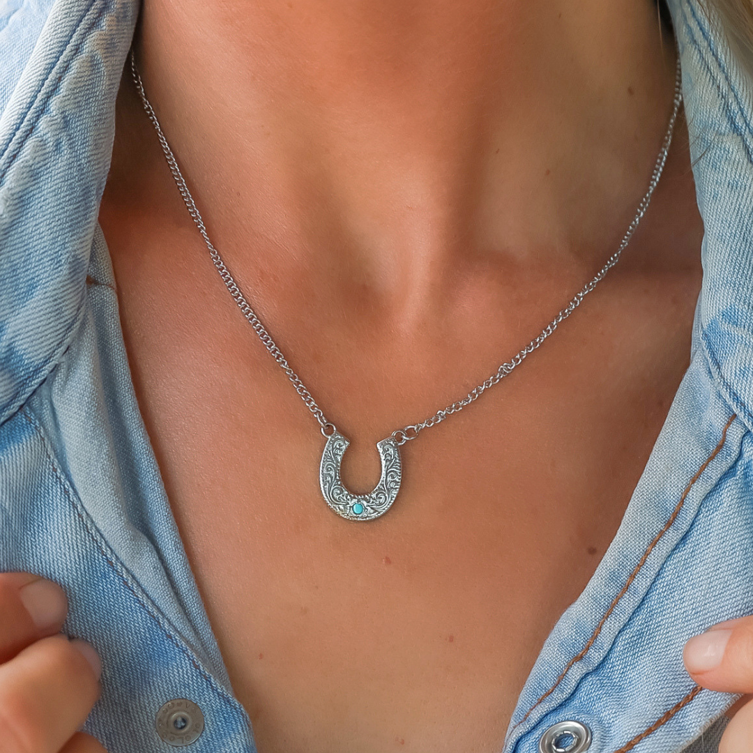 A person wearing a denim shirt showcases The Lucky Necklace—a silver horseshoe necklace with intricate detailing—elegantly paired with a turquoise pendant, shown in close-up on their neck and chest.