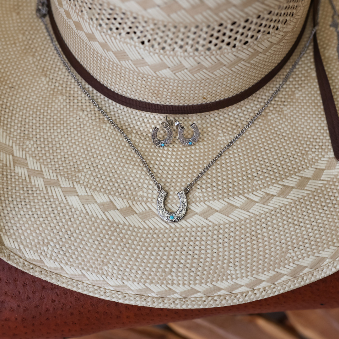 The Lucky Necklace, a silver horseshoe pendant adorned with vibrant blue stones, is showcased alongside matching earrings on a woven straw hat with a brown leather band, highlighting the elegance of this stainless steel jewelry collection.