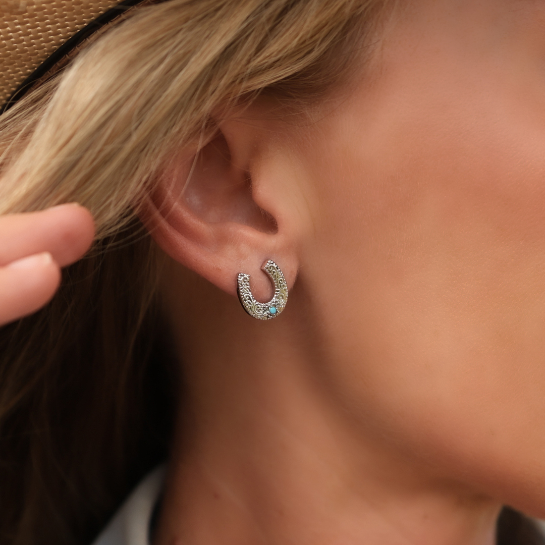 A close-up of a woman's ear wearing The Lucky Studs—small horseshoe stud earrings adorned with delicate stones. She is touching her hair and wearing a hat, highlighting the subtle elegance of this Western-inspired jewelry.