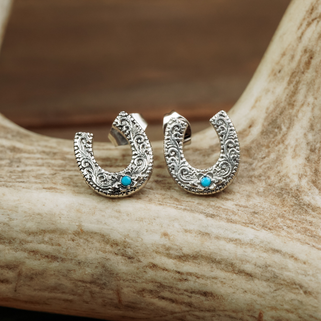 Introducing The Lucky Studs: a pair of ornate silver horseshoe-shaped earrings adorned with intricate patterns and a small turquoise stone, elegantly displayed on a light-colored antler—these striking turquoise earrings capture the timeless charm of Western jewelry.