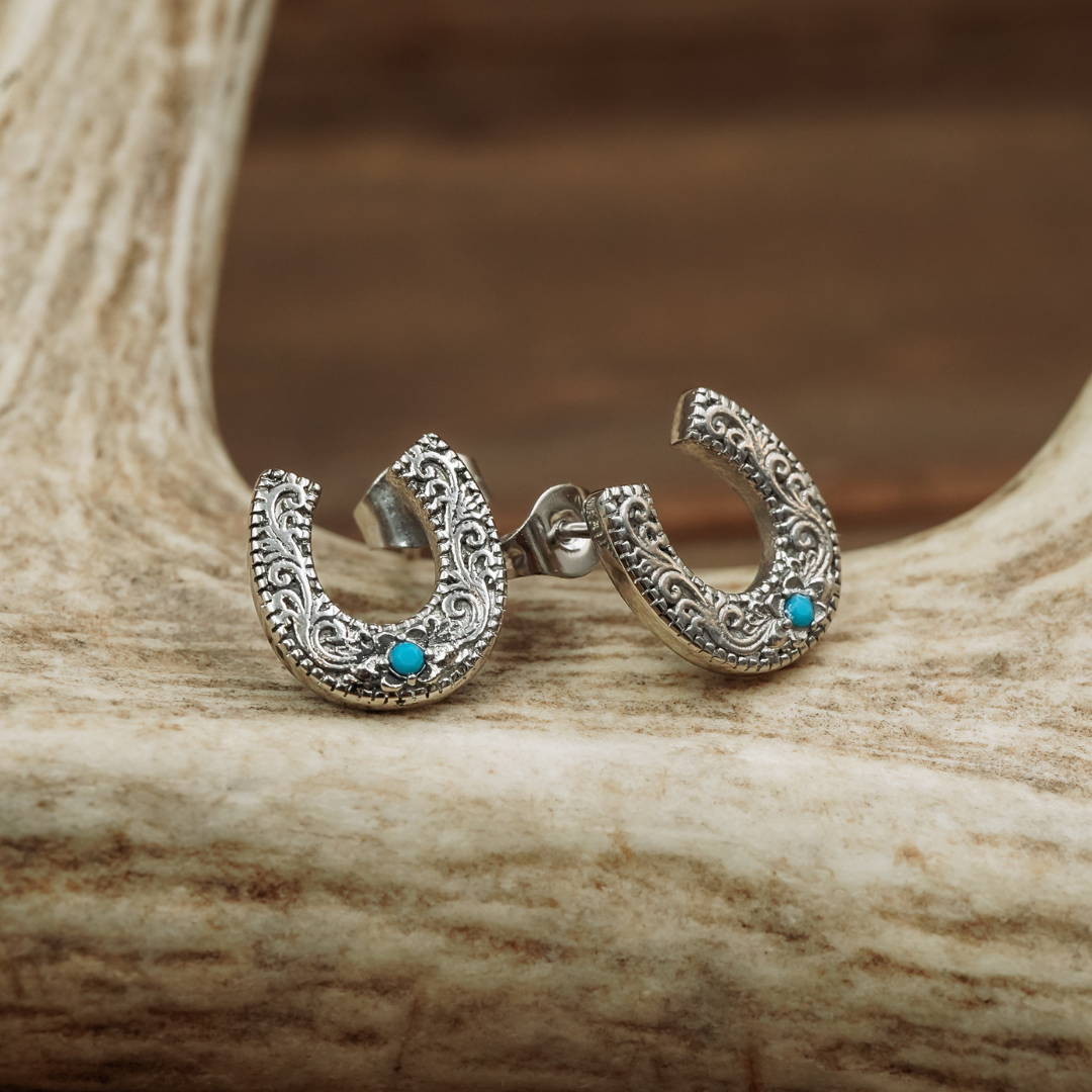 Introducing The Lucky Studs: a pair of ornate silver horseshoe stud earrings adorned with turquoise stones, beautifully displayed on a light, textured antler with a softly blurred brown background—an eye-catching piece of Western jewelry.