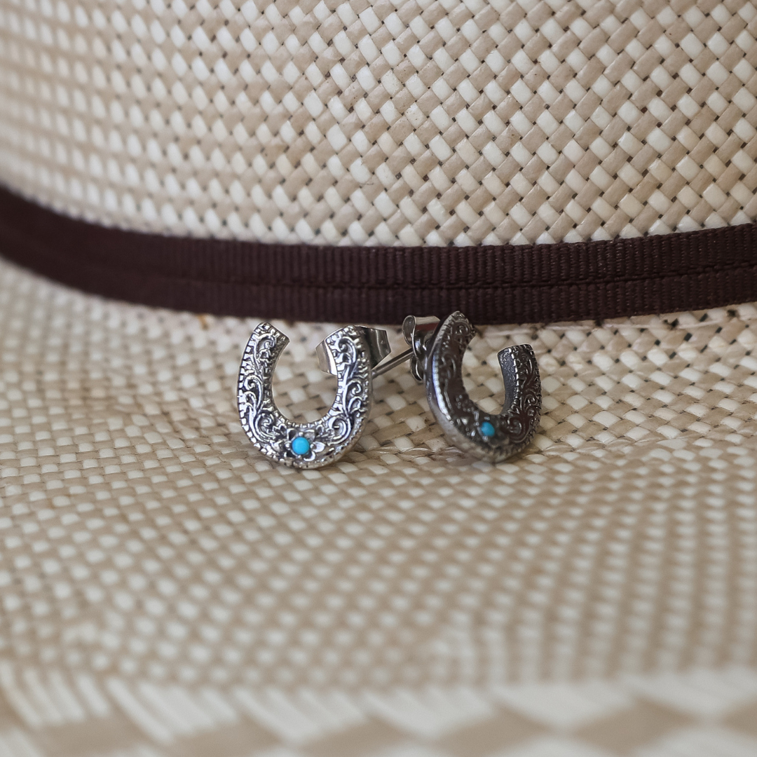 A pair of The Lucky Studs—silver horseshoe earrings accented with turquoise—rests on a woven straw hat with a brown band, capturing the timeless charm of Western jewelry.