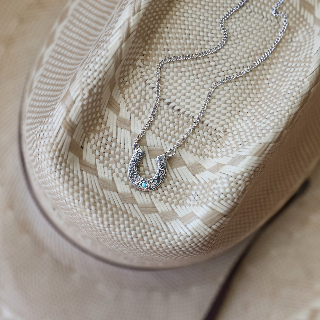 The Lucky Necklace—a silver horseshoe pendant adorned with vibrant turquoise—rests atop a beige woven cowboy hat, seamlessly blending western charm with the timeless elegance of stainless steel jewelry.