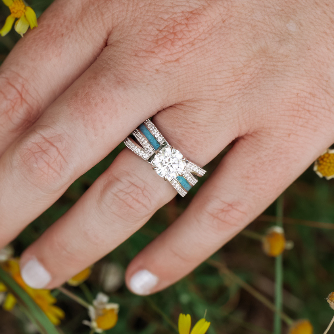 A close-up of a hand with light skin wearing the Lux Grand Chelly moissanite engagement ring with blue accents, surrounded by small yellow flowers and green grass. The nails are painted white.