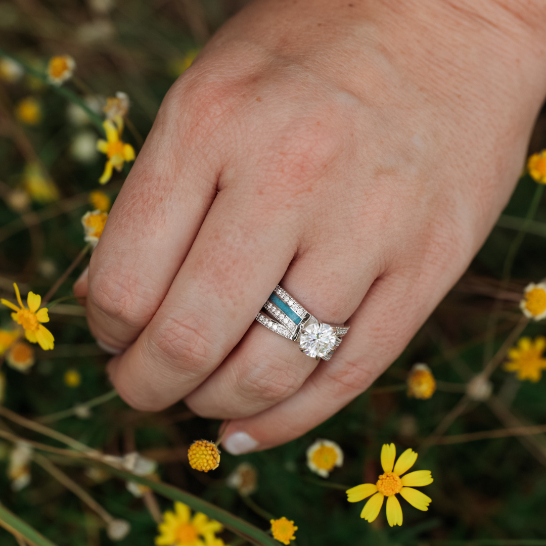 A hand wearing the Lux Grand Chelly moissanite engagement ring and wedding band rests among small yellow wildflowers.