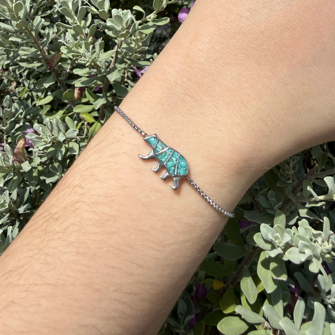 The Mama Bear Bracelet, featuring a silver band and a turquoise bear charm, is shown worn on a wrist. Green, leafy plants and gentle sunlight in the background create a serene setting—perfect for an unforgettable Mother’s Day gift.