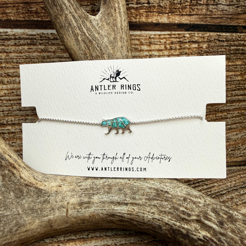 The Mama Bear Bracelet, featuring a silver finish with turquoise inlay, is displayed on a branded Antler Rings card set on rustic wood with a deer antler in the foreground—a perfect Mother’s Day gift for the special woman in your life.