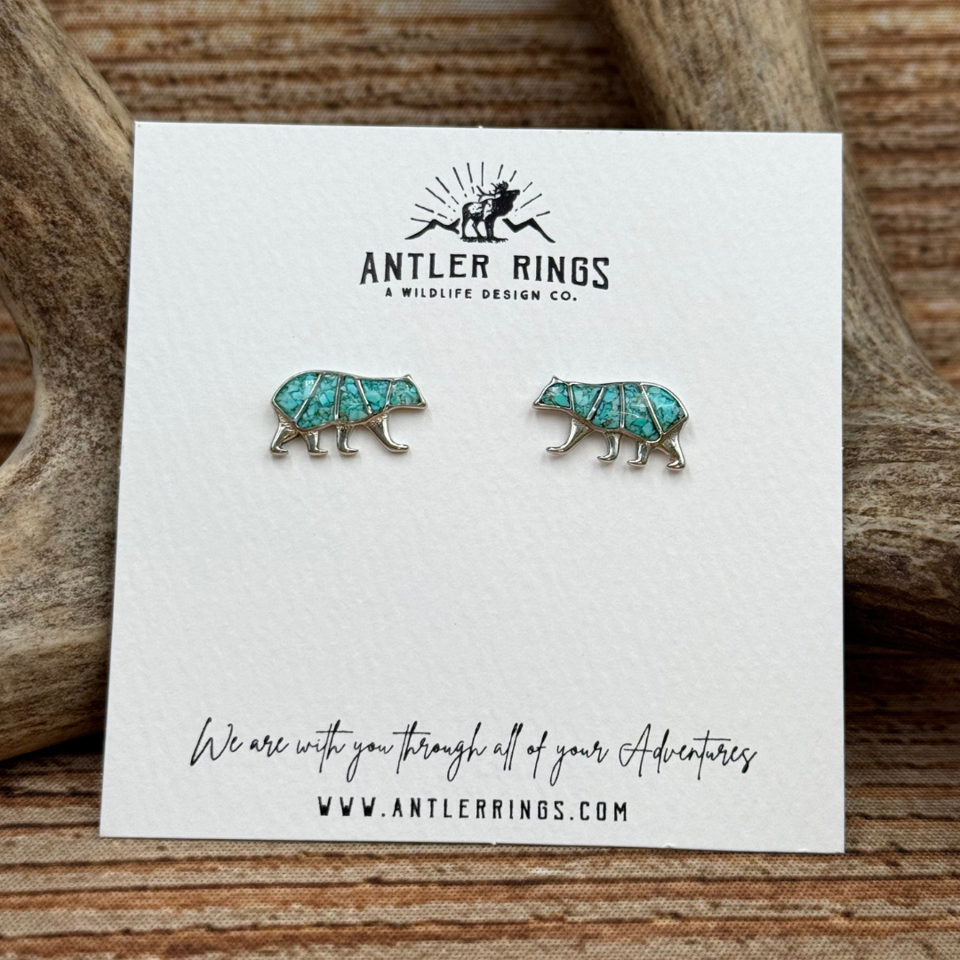 A pair of Mama Bear Stud Earrings in turquoise, featuring a charming bear silhouette design with antlers and a textured background, displayed on a branded Antler Rings card.