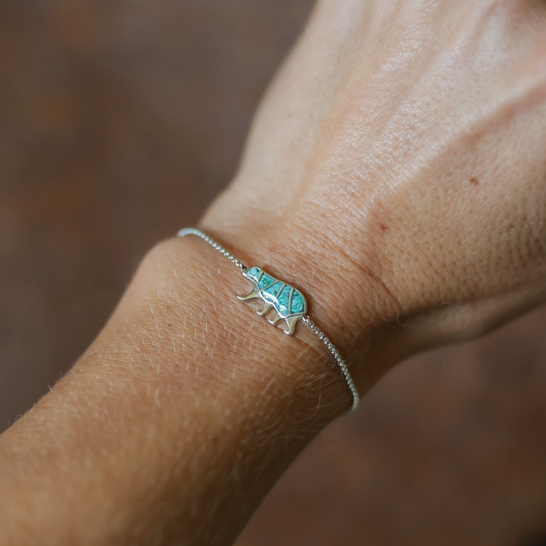 The Mama Bear Bracelet, featuring a turquoise chameleon charm on a silver band, is worn on a light-toned wrist and beautifully embodies the essence of motherhood jewelry. Set against a softly blurred brown background, this unique piece makes a thoughtful and meaningful gift.