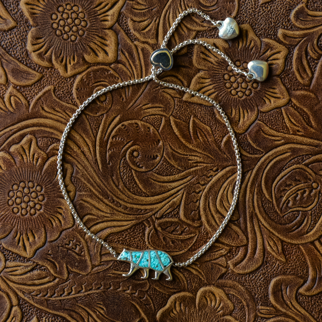 Introducing The Mama Bear Bracelet: a silver bracelet with a turquoise bear charm resting on brown, floral-embossed leather. Designed as a symbol of motherhood, it features an adjustable chain with heart-shaped ends — the perfect gift for moms who embody strength and love.