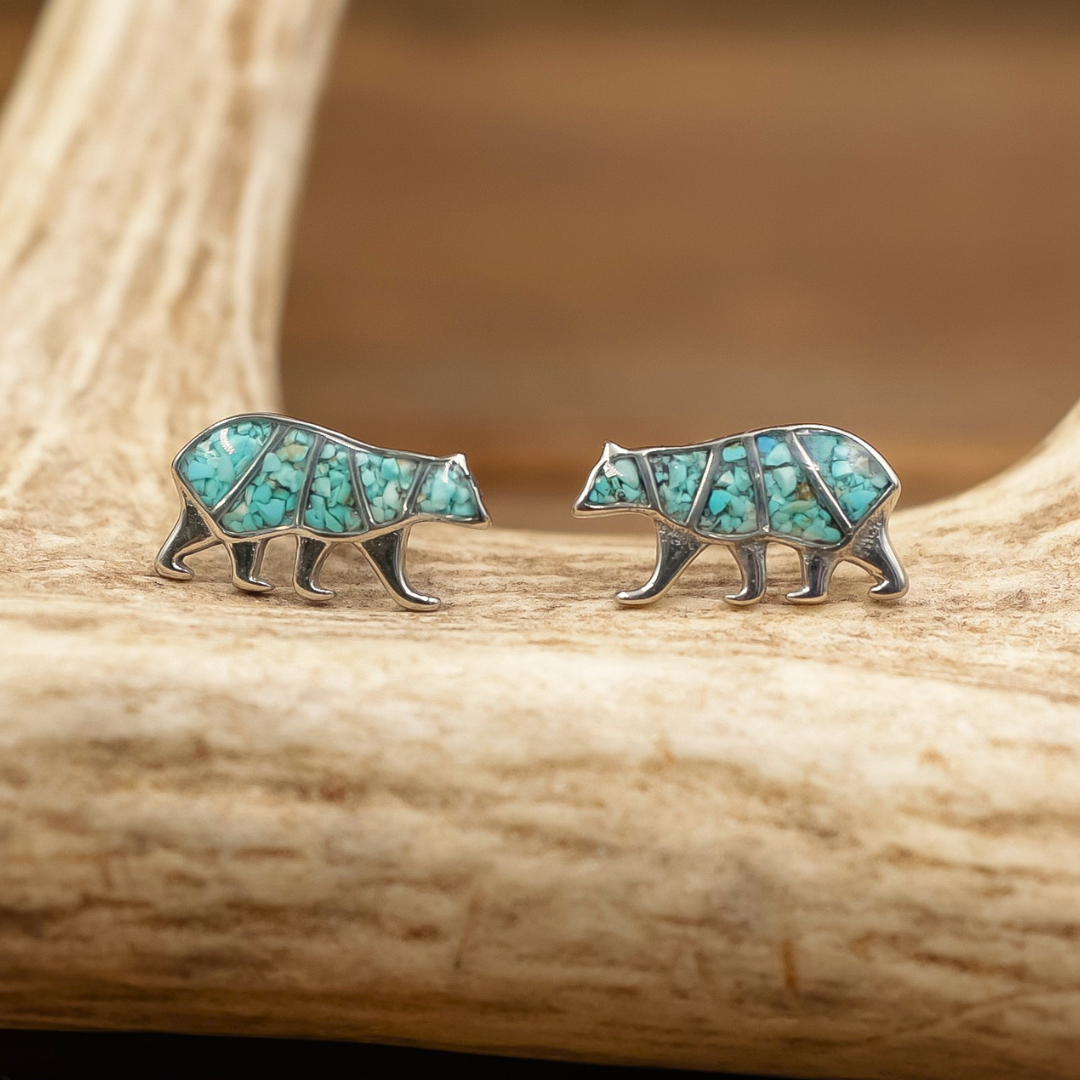 Two Mama Bear Stud Earrings, crafted in a bear shape and set with geometric turquoise inlays, rest on a light-colored wooden surface. These earrings make a beautiful piece of motherhood jewelry.