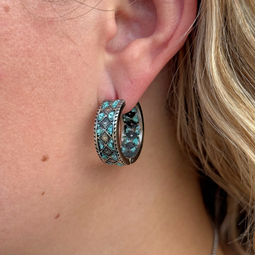 A close-up of a person’s ear wearing The Meggan Hoops highlights the bold, silver and turquoise patterned design that exemplifies the unique style of Western jewelry. The person has light skin and blonde hair.
