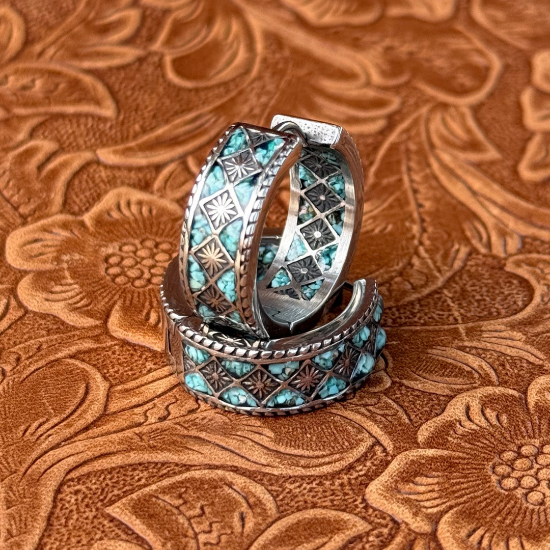 The Meggan Hoops—silver rings adorned with turquoise and engraved geometric patterns—are displayed upright on an embossed brown leather surface with floral designs, capturing the timeless elegance of Western jewelry.