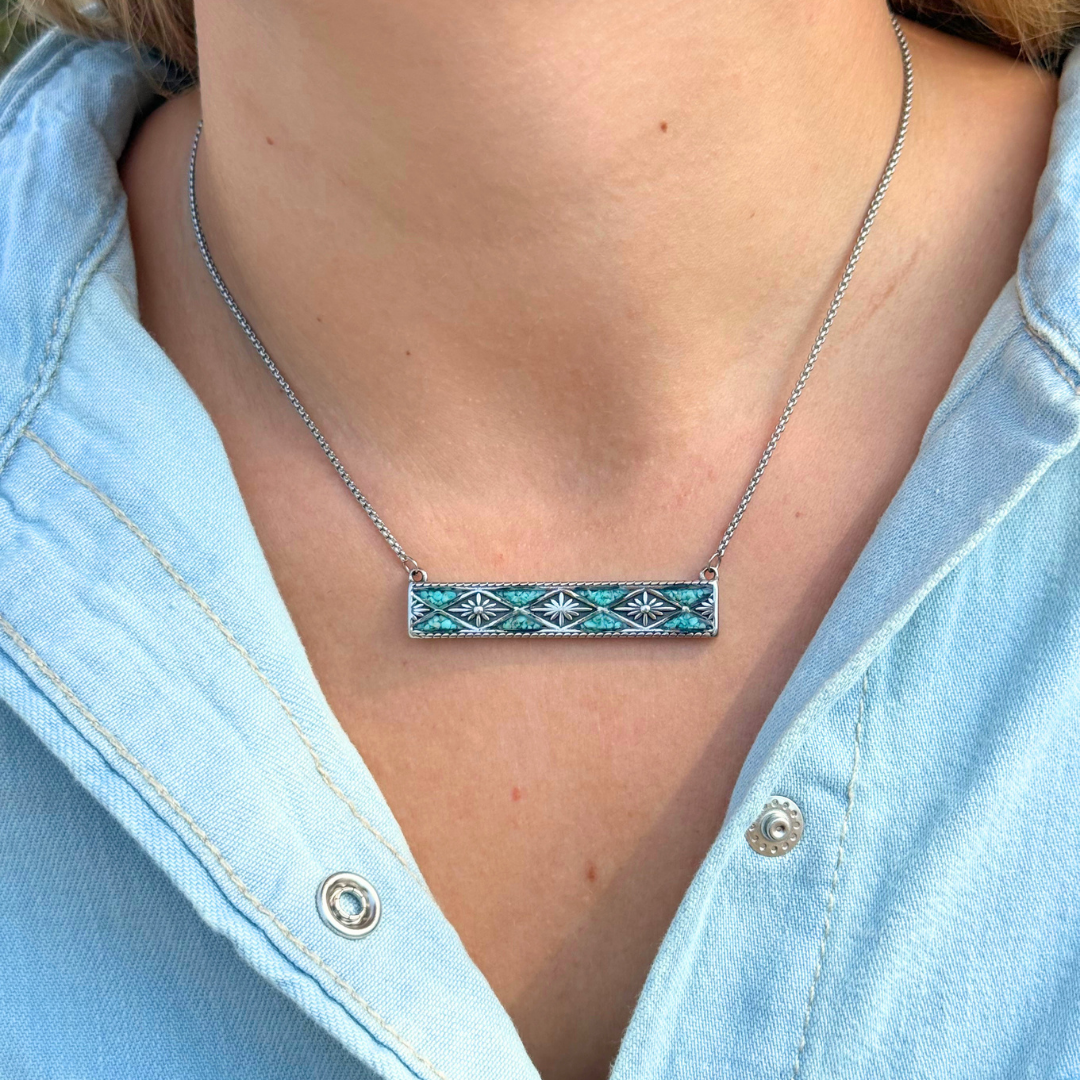 A close-up of a person wearing The Meggan, a silver bar necklace with a rectangular pendant featuring a blue and black geometric pattern inspired by Western jewelry, over a light denim shirt.