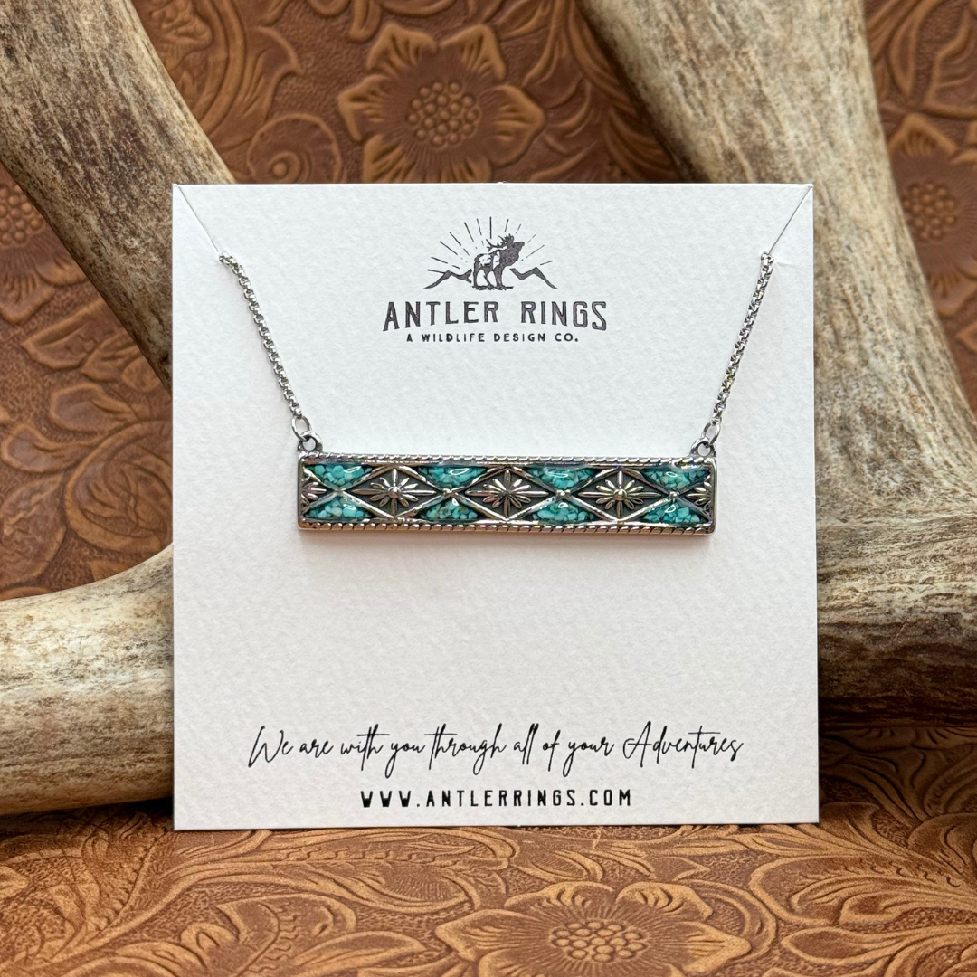 The Meggan, a silver bar necklace featuring crushed turquoise inlay, is displayed on an Antler Rings card set before an antler. Ideal for fans of Western jewelry, the card showcases the company name, logo, website, and an encouraging message for adventurers.