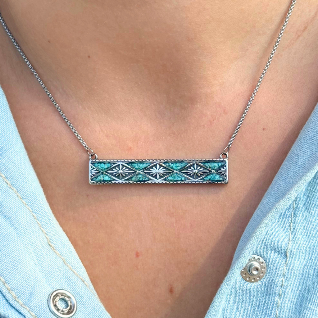 A close-up of a person wearing The Meggan, a silver Western jewelry bar necklace featuring a rectangular pendant with crushed turquoise and white geometric detailing, paired with a light blue denim shirt.