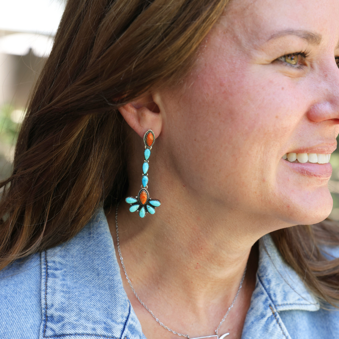 A woman with long brown hair, wearing a light denim jacket, smiles while showing off The Miranda Drop Earrings—statement earrings featuring turquoise and orange stones that make a striking example of Western jewelry.