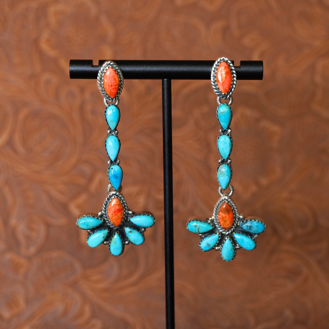 The Miranda Drop Earrings feature long, dangling statement drops with Mojave turquoise and red-orange stones set in silver, showcased on a black stand against a richly patterned brown background—a striking example of Western jewelry.