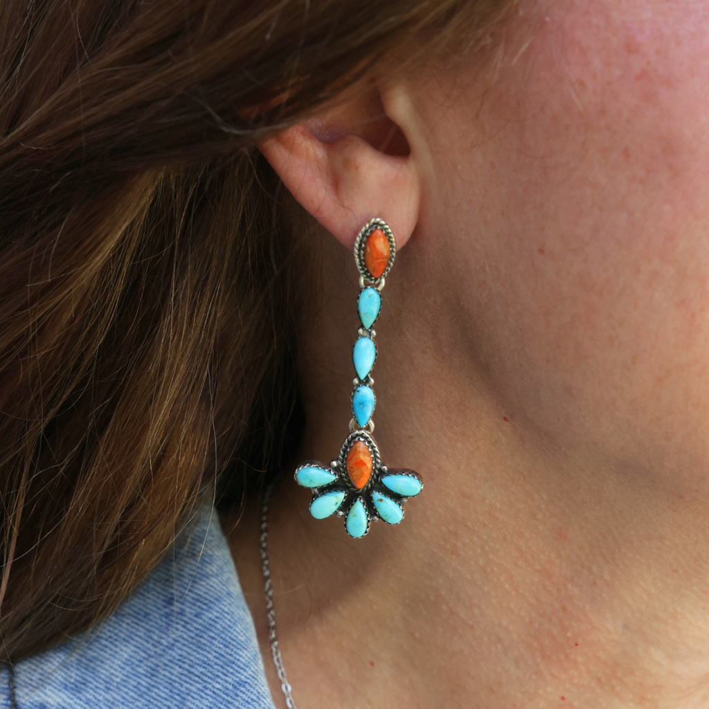 A close-up of a person wearing The Miranda Drop Earrings—long Mojave turquoise and orange stone earrings set in silver, featuring a floral-inspired design. Part of their brown hair, ear, and denim jacket are visible, highlighting these eye-catching statement pieces that reflect the beauty of Western jewelry.