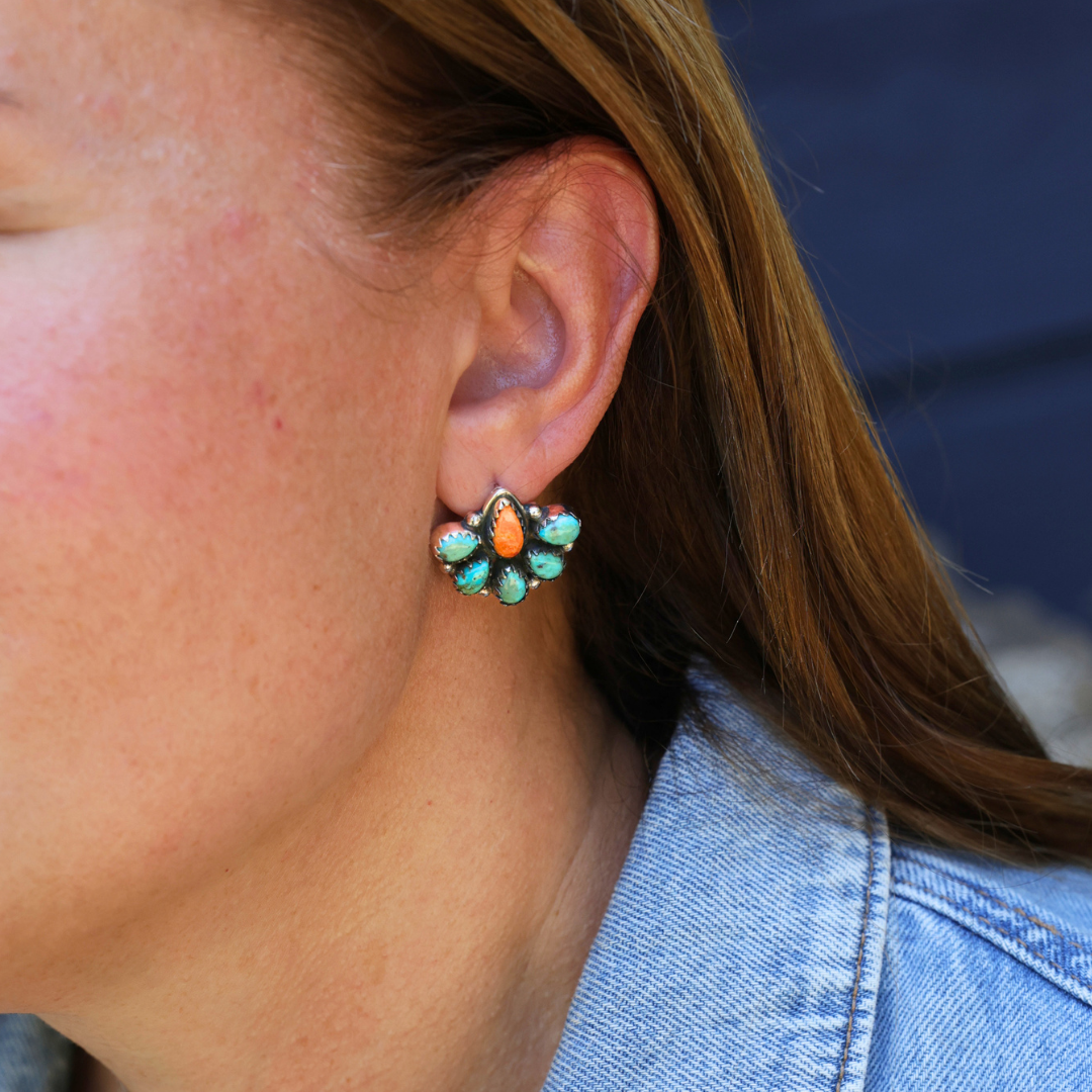 A close-up of a person’s ear wearing The Miranda Stud Earrings, featuring a floral design with Mojave turquoise and orange stones. The person has light skin, brown hair, and is dressed in a light blue denim jacket.