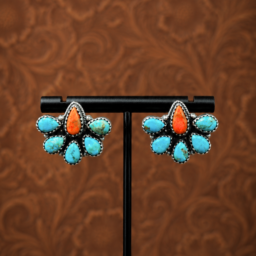 The Miranda Stud Earrings feature silver studs adorned with orange coral and Mojave turquoise stones arranged in a floral pattern, displayed on a black stand against a brown ornate background.
