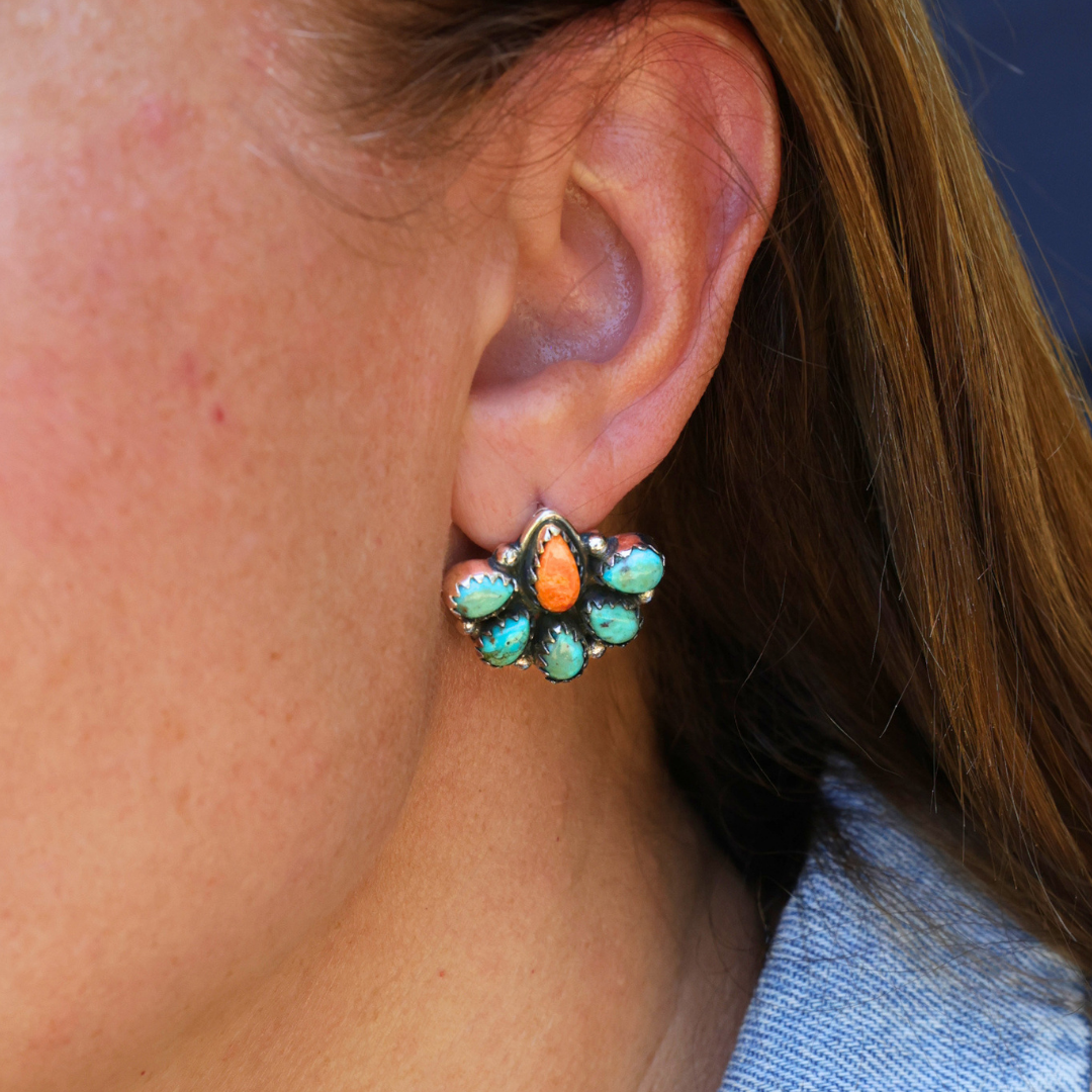 A close-up of a person’s ear wearing The Miranda Stud Earrings—flower-shaped studs adorned with five vibrant stones, including orange and Mojave turquoise, set in silver. The person has light brown hair and is dressed in a light blue top.