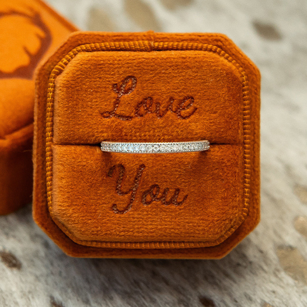 A delicate Moissanite Eternity Stacking Band, crafted from 925 Sterling Silver and adorned with a thin line of sparkling moissanites, rests in an orange velvet box with “Love You” embroidered inside, placed on a speckled surface.