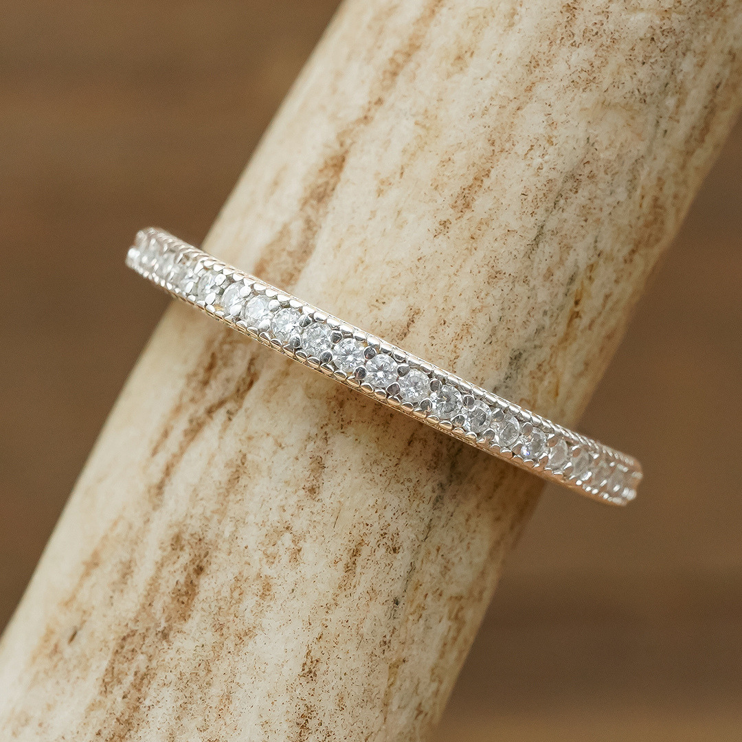 The Moissanite Eternity Stacking Band, a slender gold ring adorned with a row of brilliant moissanite stones, is displayed on a textured, light-colored surface.