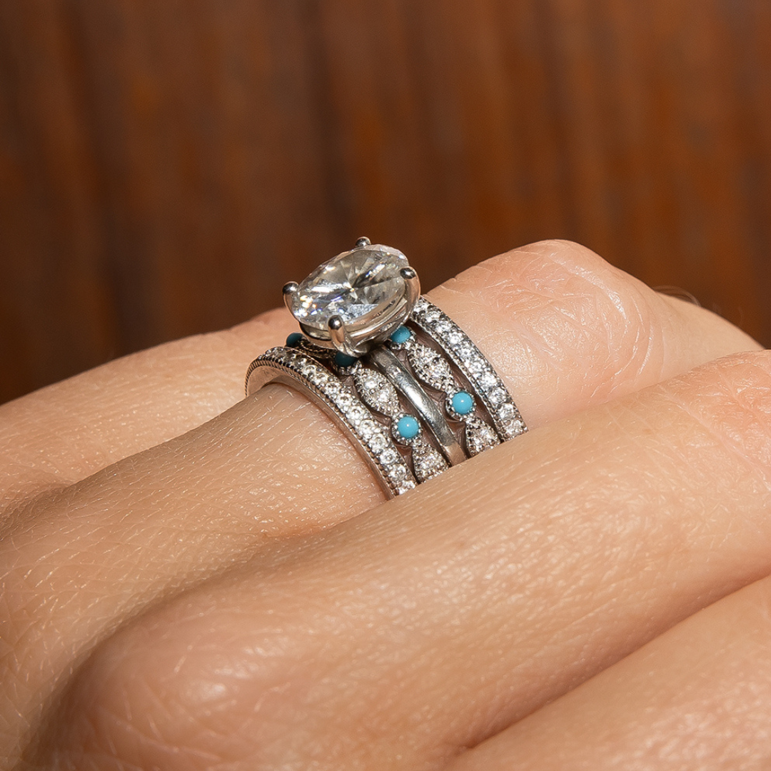 A close-up of a hand wearing three stacked silver rings with diamonds and small turquoise stones, featuring a large central clear gemstone. One ring is the sparkling Moissanite Eternity Stacking Band, while another is crafted from 925 Sterling Silver—each stacking ring brings its own unique charm to the elegant ensemble.