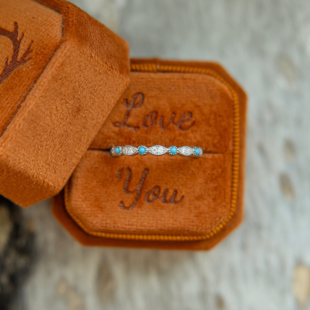 The Moissanite Turquoise Vintage Stacking Band is a delicate sterling silver ring adorned with turquoise and sparkling moissanite stones, presented in a brown suede box featuring “Love You” embroidered inside the lid.