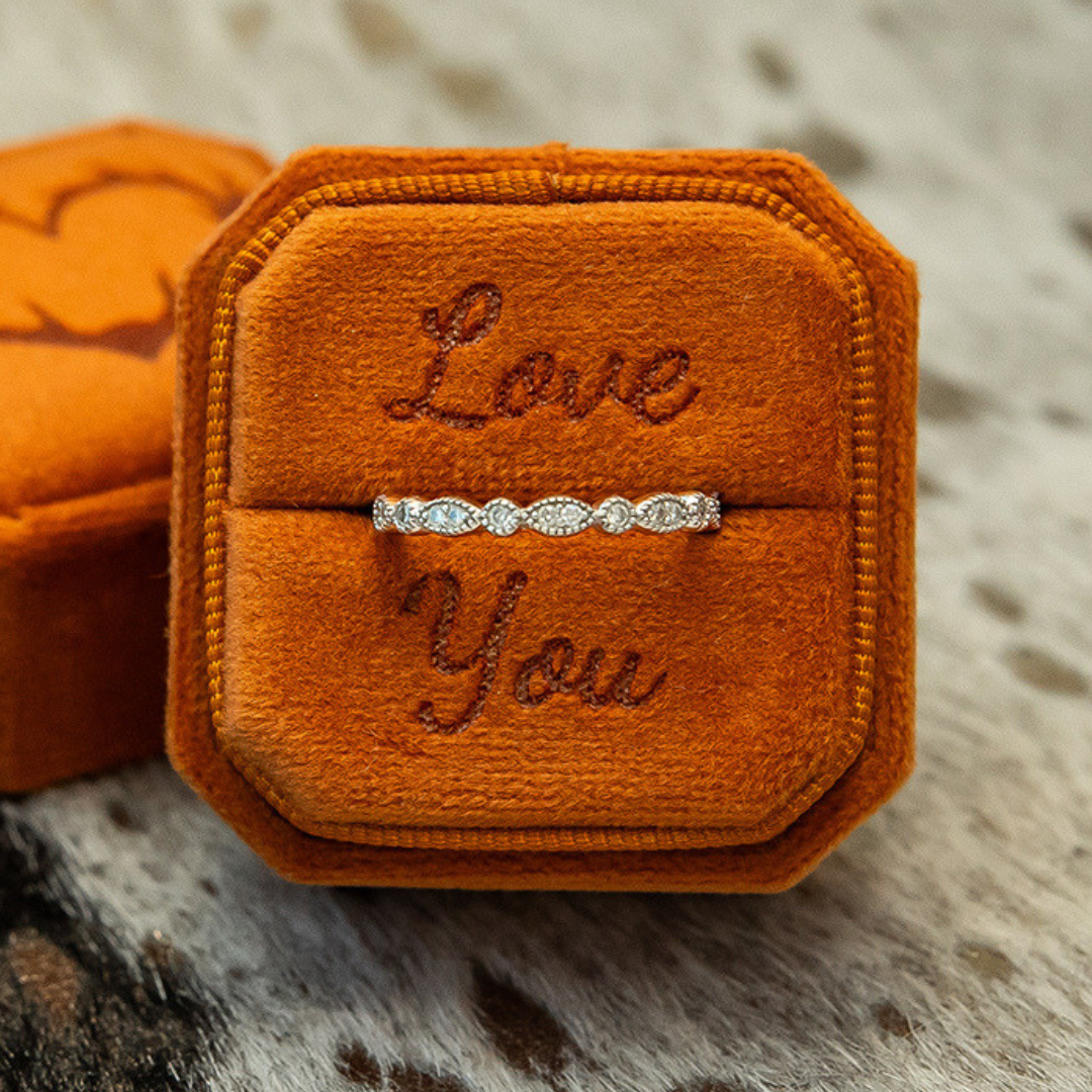 A Moissanite Vintage Stacking Band rests in an orange velvet box, with “Love You” embroidered on the lid, placed on a soft, textured surface.