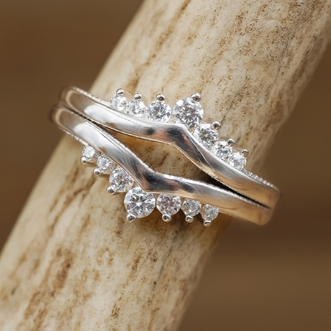 A sterling silver ring with a wavy design and two rows of round, clear gemstones rests on a textured, light brown surface, making it a stunning addition to the Moissanite Wishbone Stacker Set.