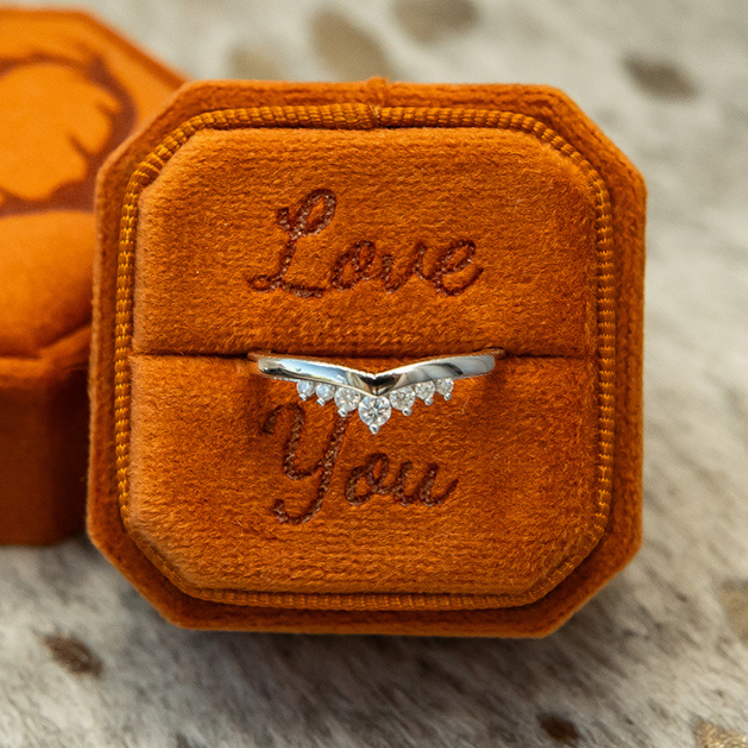 A Moissanite Wishbone Stacking Band in a brown velvet box embroidered with the words “Love You” sits on a soft, light-colored surface.