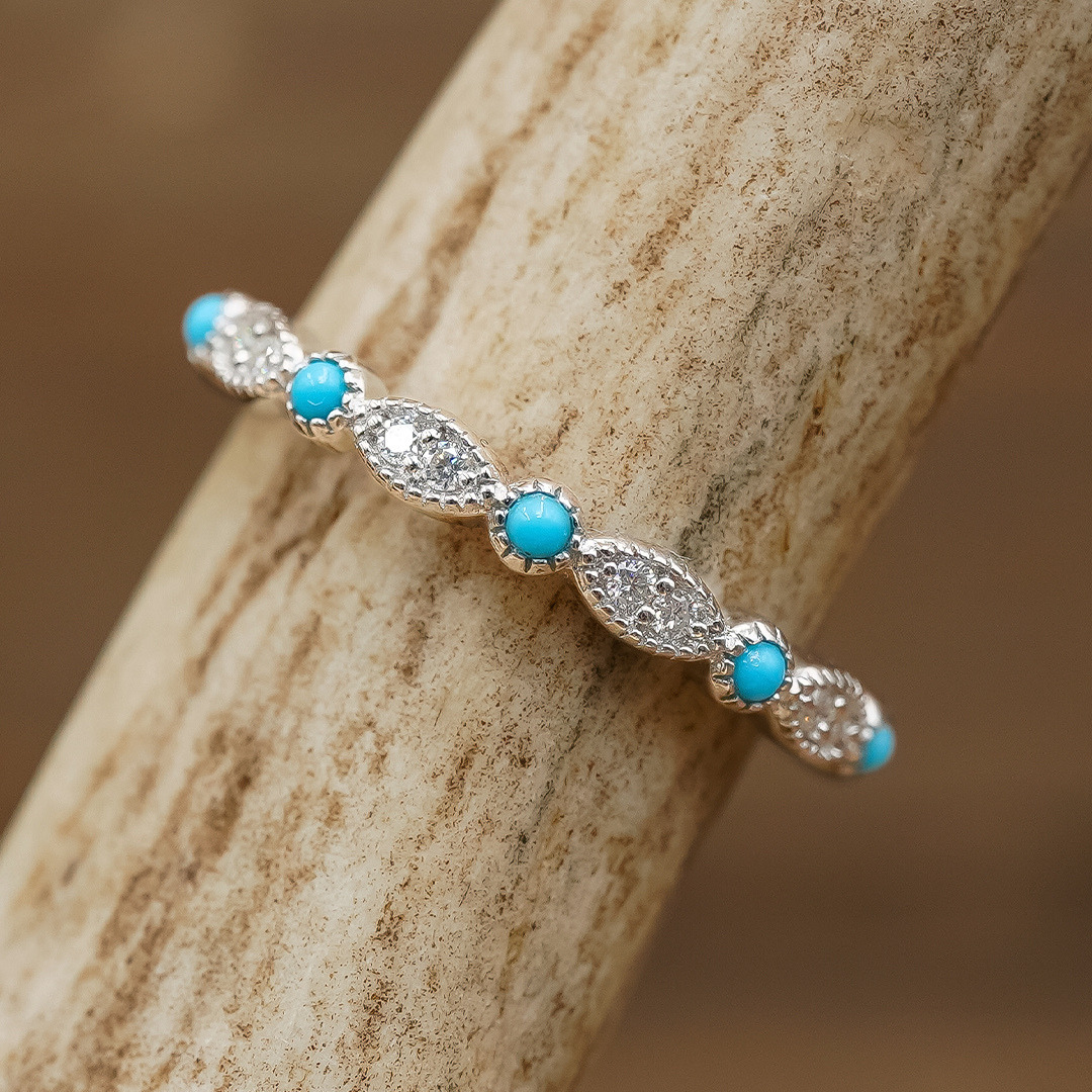 The Moissanite Turquoise Vintage Stacking Band features alternating small turquoise stones and clear oval moissanite crystals, beautifully displayed on a textured light brown surface.