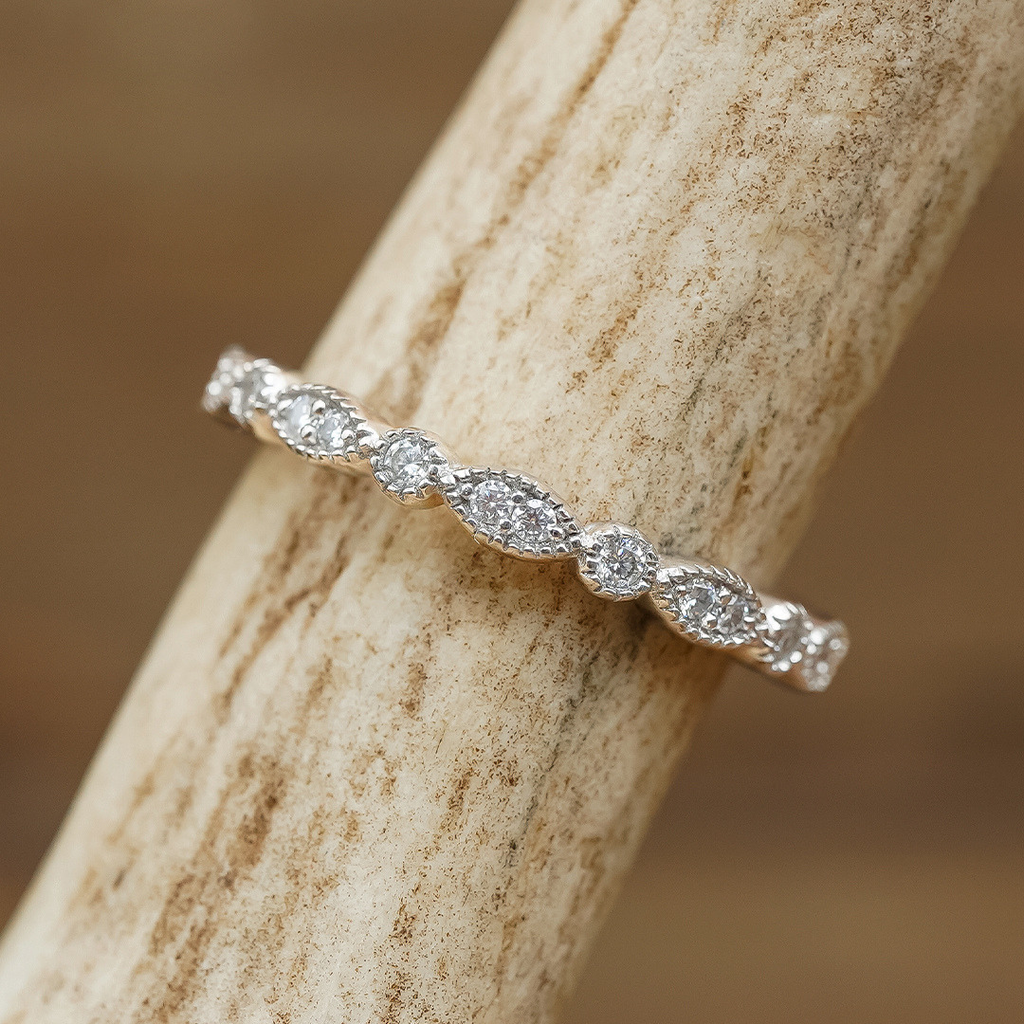 The Moissanite Vintage Stacking Band, featuring alternating round and marquise-shaped stones in a delicate gold setting and evoking vintage elegance, rests on a textured, light-colored surface.