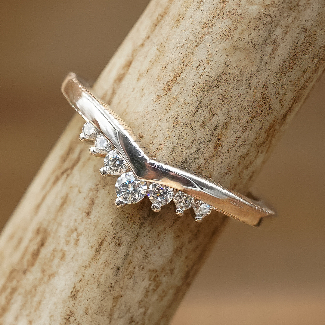 The Moissanite Wishbone Stacking Band, crafted in sterling silver with a V-shaped design and five small round-cut moissanite stones set along the curve, is displayed on a light-colored textured surface.